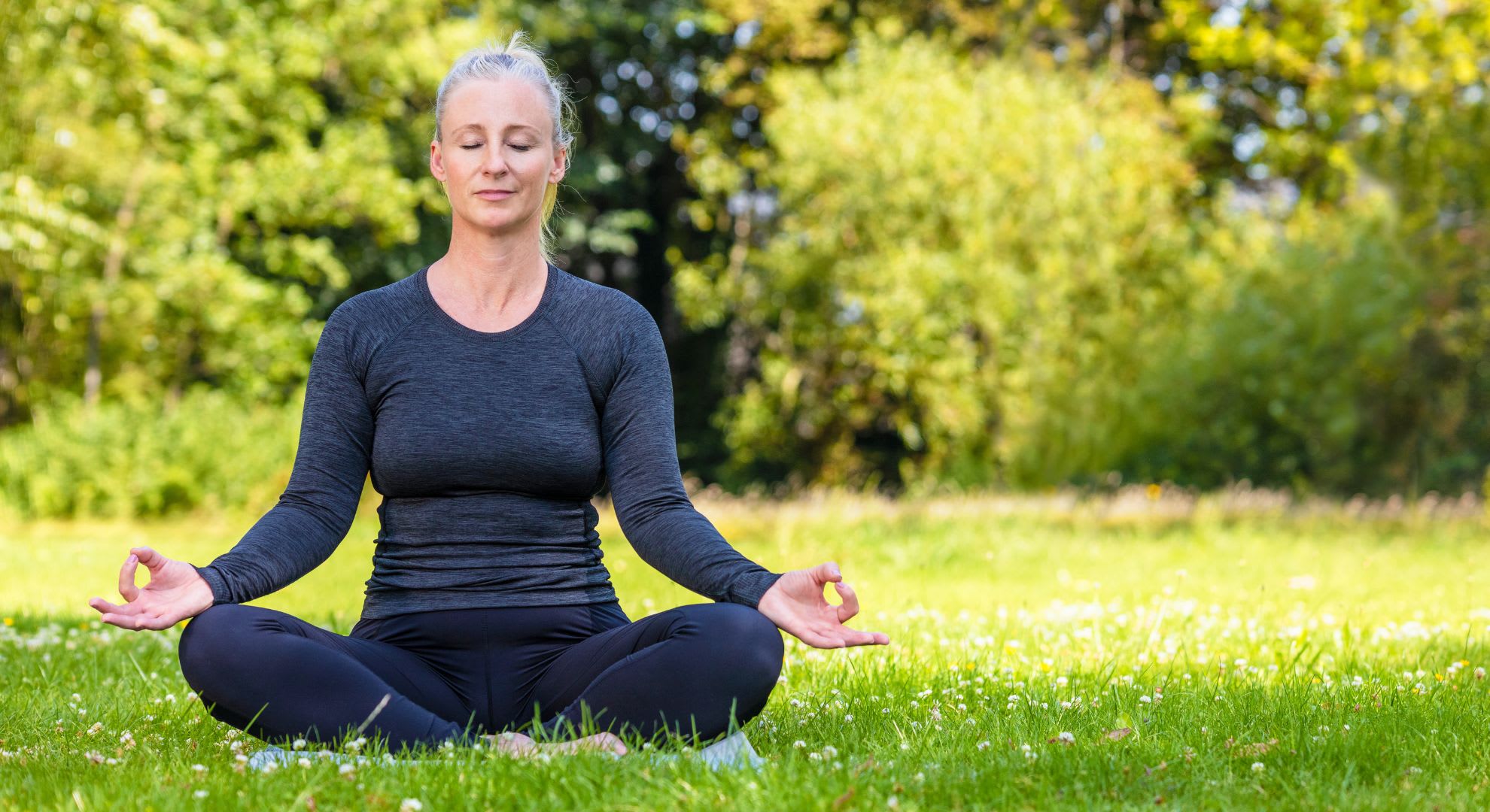 A person with grey hair is meditating in a seated position on grass in a park, embracing tranquility akin to a house call doctor bringing calm through care. With eyes closed and hands in a mudra gesture, trees stand serenely in the background.
